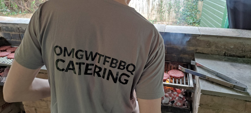 A person wearing an OMGWTFBBQ Catering t-shirt standing in front of a BBQ. Various uncooked burgers are visible.